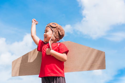 Happy Asian funny child or kid little boy smile wear pilot hat and goggles play toy cardboard airplane wing flying raises hand up against summer blue sky cloud background, Startup freedom concept