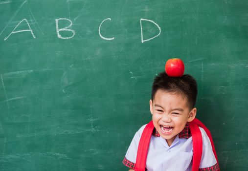 Back to School. Happy Asian funny cute little child boy from kindergarten in student uniform with school bag and red apple on head smiling on green school blackboard, First time to school education