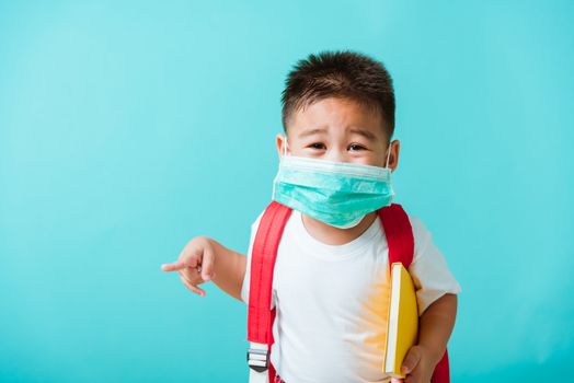 Back to school coronavirus Covid-19 education. Portrait Asian little child boy kindergarten wear face mask protective and school bag hold book before going to school pointing to side away, studio shot