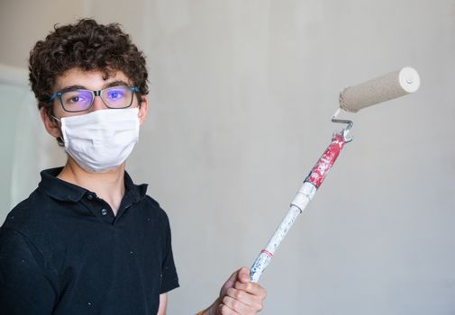 Renovate the walls of the house by painting them in the time of the coronavirus: the Caucasian boy has a mask to protect himself from the virus. Pass the impregnated roller of gray color on the wall.