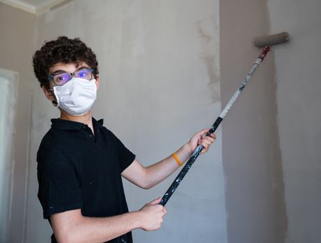 Renovate the walls of the house by painting them in the time of the coronavirus: the Caucasian boy has a mask to protect himself from the virus. Pass the impregnated roller of gray color on the wall.