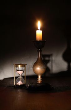 Elegant brass candlestick with lighting candle near hourglass on dark background