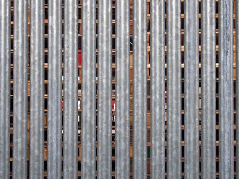 a vertical galvanized steel fence with wooden pallets in outdoor storage visible between the gaps