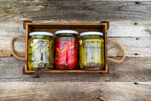 Glass jars with pickled red bell peppers and pickled cucumbers (pickles) isolated in wooden crate. Jars with variety of pickled vegetables. Preserved food concept in a rustic composition.