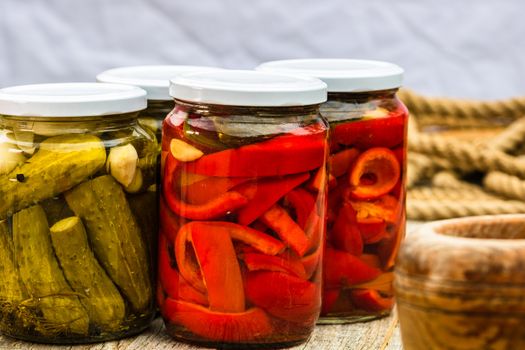 Glass jars with pickled red bell peppers and pickled cucumbers (pickles) isolated. Jars with variety of pickled vegetables. Preserved food concept in a rustic composition.