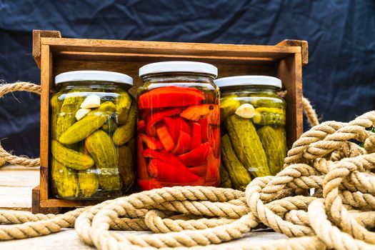 Glass jars with pickled red bell peppers and pickled cucumbers (pickles) isolated in wooden crate. Jars with variety of pickled vegetables. Preserved food concept in a rustic composition.