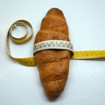 soft measuring ruler wrapped around a croissant as a symbol of unhealthy nutrition, on a white background