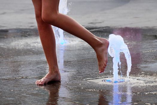 a girl touches a fountain on the sidewalk with her bare foot