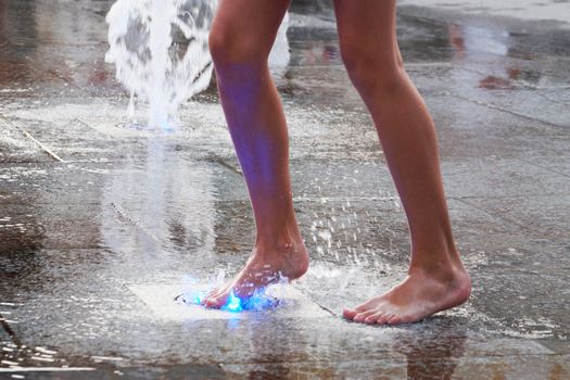 a girl touches a fountain on the sidewalk with her bare foot