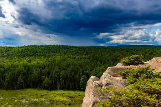 A cloudy sky stretches off towards the horizon with distant rain falling above a forest of green trees. The woods are seen from above with a rocky cliff in the foreground.
