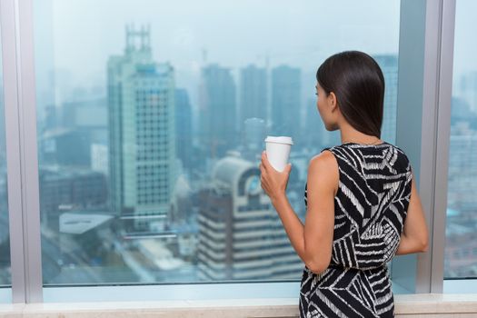 Business woman drinking coffee in office during lunch break looking at city's skyline view from window thinking, Shanghai, China.