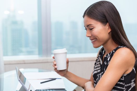Businesswoman drinking coffee using laptop at office desk. Asian woman working on computer online at home.