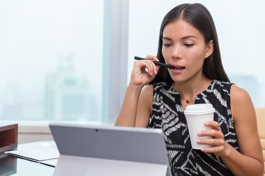 Serious Asian business woman busy working on laptop at work or home office reading and taking notes while drinking coffee. Beautiful multiracial businesswoman being concentrated.