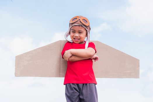 Happy Asian funny child or kid little boy smile wear pilot hat and goggles play toy cardboard airplane wing flying stand crossed arm against summer blue sky cloud background, Startup freedom concept