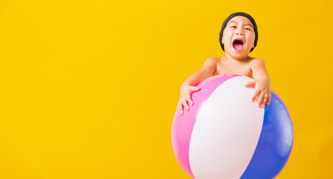 Summer vacation concept, Portrait Asian happy cute little child boy smiling in swimsuit hold beach ball, Kid having fun with inflatable ball in summer vacation, studio shot isolated yellow background