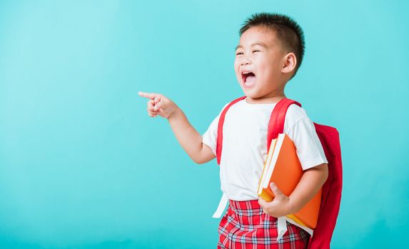 Back to school concept. Portrait Asian happy funny cute little child boy smile hug books and point finger to side away space, isolated blue background. Kid from preschool kindergarten with school bag