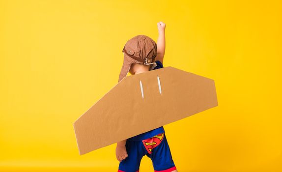 Back of Happy Asian handsome funny child or kid little boy smile wear pilot hat play raise hand up with toy cardboard airplane wings flying, studio shot isolated yellow background, Startup freedom