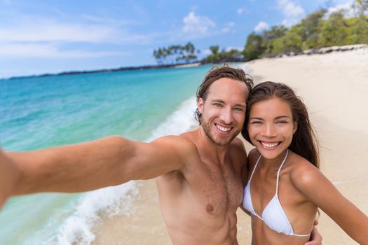 Happy seflie couple taking picture with smartphone on beach vacation smiling at camera. Asian woman, Caucasian man. Young multiracial people having fun together in summer travel destination.