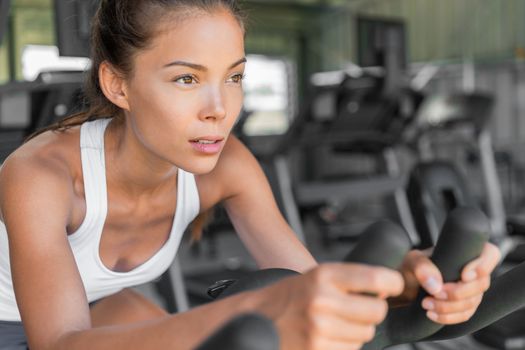 Girl doing cardio class at gym on exercise bike. Indoor cycling with wheels spinning. Focus and weight loss motivation. Young mixed race Asian Caucasian woman training fitness workout on bicycle.