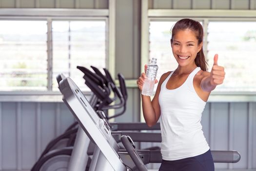 Happy gym woman doing thumbs up during cardio workout on treadmill machine drinking water. Health and fitness.