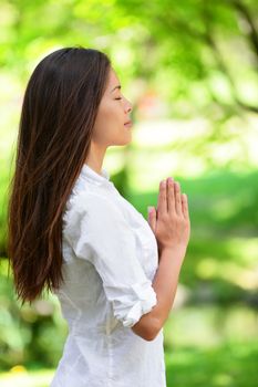 Young woman with hands clasped meditating in park. Side view of beautiful woman in casuals. Attractive female with eyes closed practicing yoga in nature.