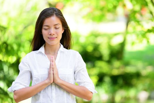Attractive young woman meditating in park. Beautiful woman in casuals is standing with hands clasped. Young female with eyes closed practicing yoga in nature.
