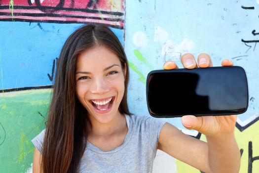 Happy young woman showing blank smartphone screen. Cheerful female is holding black mobile phone outdoors. The empty space on cellphone can be used for advertisement purpose.