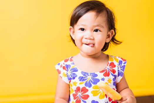 Happy portrait Asian baby or kid cute little girl attractive laugh smile wearing dick pattern shirt holds and eating sweet wooden ice cream, studio shot isolated on yellow background, summer concept