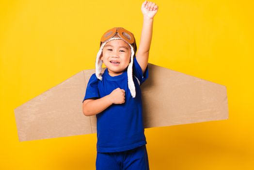 Happy Asian handsome funny child or kid little boy smile wear pilot hat play and goggles raise hand up with toy cardboard airplane wings flying, studio shot isolated yellow background, Startup freedom