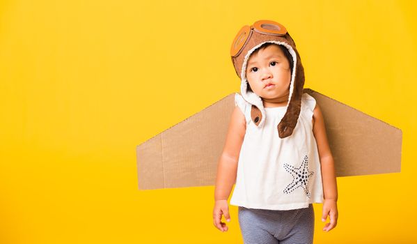 Happy Asian beautiful funny baby little girl smile wear pilot hat playing and goggles with toy cardboard airplane wings flying, studio shot isolated yellow background, Startup freedom concept