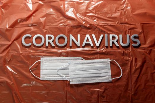 the word coronavirus laid with silver letters on crumpled orange plastic film with white medical facial masks - flat lay, centered composition and dramatic light