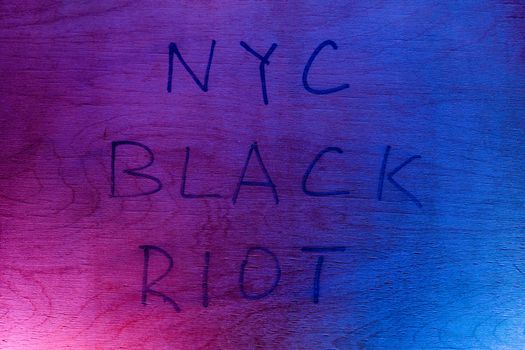 The words NYC black riot handwritten with black paint marker on flat plywood surface under red and blue police lights. Concept of night disorder in New York City.