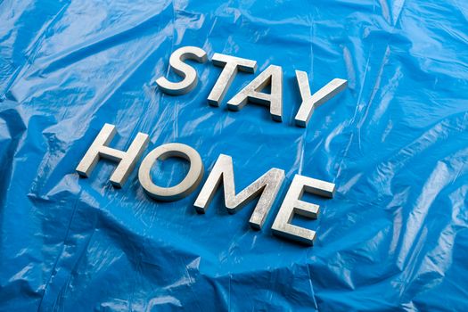 the words stay home laid with silver metal letters on crumpled blue plastic background in slanted diagonal perspective.