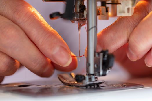 elder woman fingers inserting a red thread into a sewing machine needle, close-up with selective focus