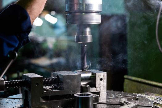 Industrial steel drilling process with smoke close-up with selective focus. Manual feed by hand. Workpiece in vise on table. High contrast with background blur.