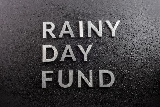the words rainy day fund laid with silver metal letters on black background, covered with water drops, flat lay perspective, directly above view