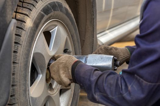 Hands of automotive mechanic unscrewing nuts with pneumatic impact wrench during car wheels season changing. Close-up view with selective focus and background blur.