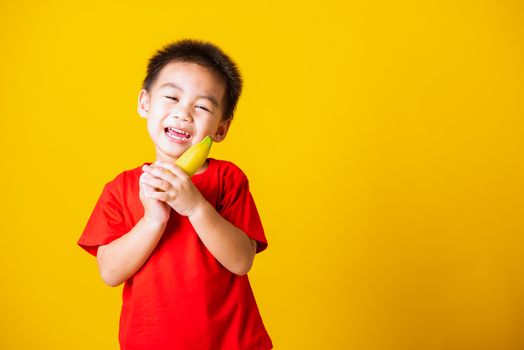 Happy portrait Asian child or kid cute little boy attractive smile wearing red t-shirt playing holds banana fruit, studio shot isolated on yellow background