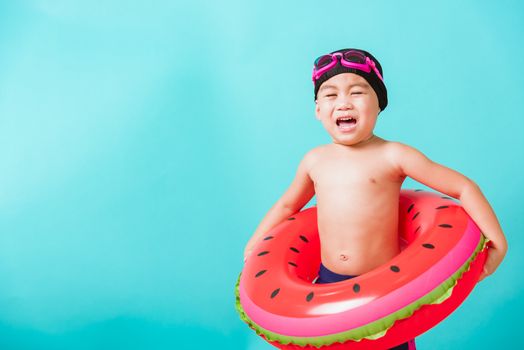 Summer vacation concept, Portrait Asian happy cute little child boy wear goggles and swimsuit hold watermelon inflatable ring, Kid having fun on summer vacation, studio shot isolated blue background