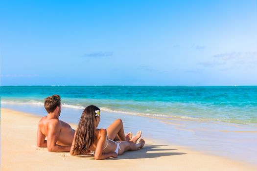 Couple on beach travel honeymoon vacation lying down sunbathing relaxing on luxury holiday in idyllic destination. Young tourists in love.