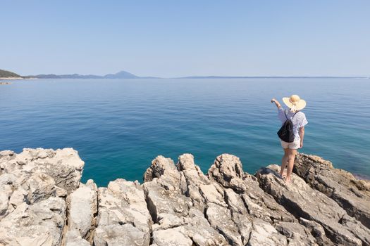 Woman traveler wearing straw summer hat and backpack, standing at edge of the rocky cliff looking and pointing at big blue sea and islands in on the horizon.