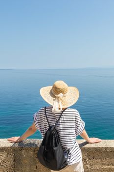Rear view of woman traveler wearing straw summer hat and backpack,leaning against a stone wall looking at big blue sea and islands in on the horizon. Copy space.