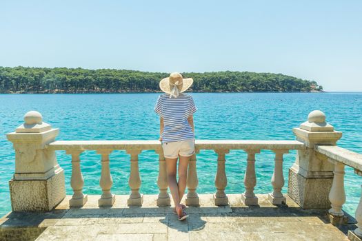 Rear view of woman wearing straw summer hat ,leaning against elegant old stone fence of coastal villa, relaxing while looking at blue Adriatic sea, on Losinj island Croatia.