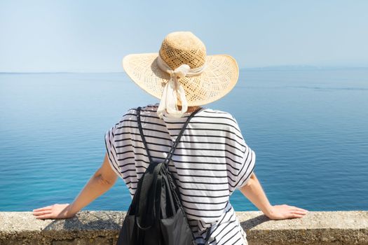 Rear view of woman traveler wearing straw summer hat and backpack,leaning against a stone wall looking at big blue sea and islands in on the horizon. Copy space.