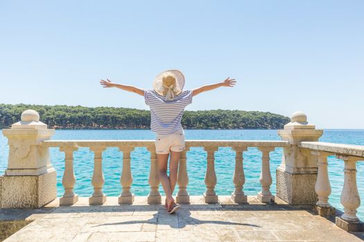 Rear view of happy woman on vacation, wearing straw summer hat ,standing on luxury elegant old stone balcony of coastal villa, relaxing, arms rised to the sun, looking at blue Adriatic sea.