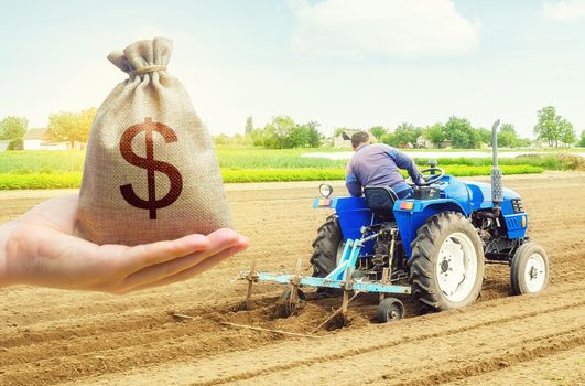 A hand holds out a dollar money bag on a background of farmer on a tractor making mounds rows on a farm field. Lending farmers for purchase land and seed, modernization. Support subsidies. Farm loans