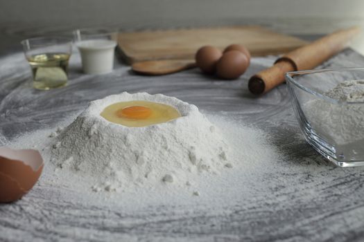 Broken chicken egg in a pile of flour, olive oil, milk, kitchen tool on gray table background. Products for baking bakery products. Cutting board, rolling pin, flour sieve, wooden spoon. bread or cake