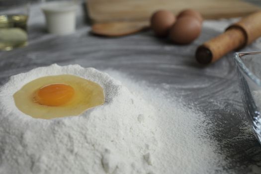 Broken chicken egg in a pile of flour, olive oil, milk, kitchen tool on gray table background. Products for baking bakery products. Cutting board, rolling pin, flour sieve, wooden spoon. bread or cake