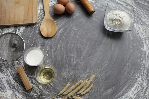 Chicken egg, flour, olive oil, milk, wheat ears, kitchen tool on gray table background. Products for baking bakery products. Cutting board, rolling pin, flour sieve, wooden spoon. For bread or cake