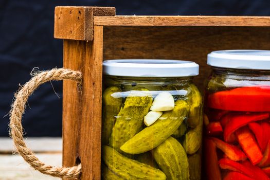 Wooden crate with glass jars with pickled red bell peppers and pickled cucumbers (pickles) isolated. Jars with variety of pickled vegetables. Preserved food concept in a rustic composition.